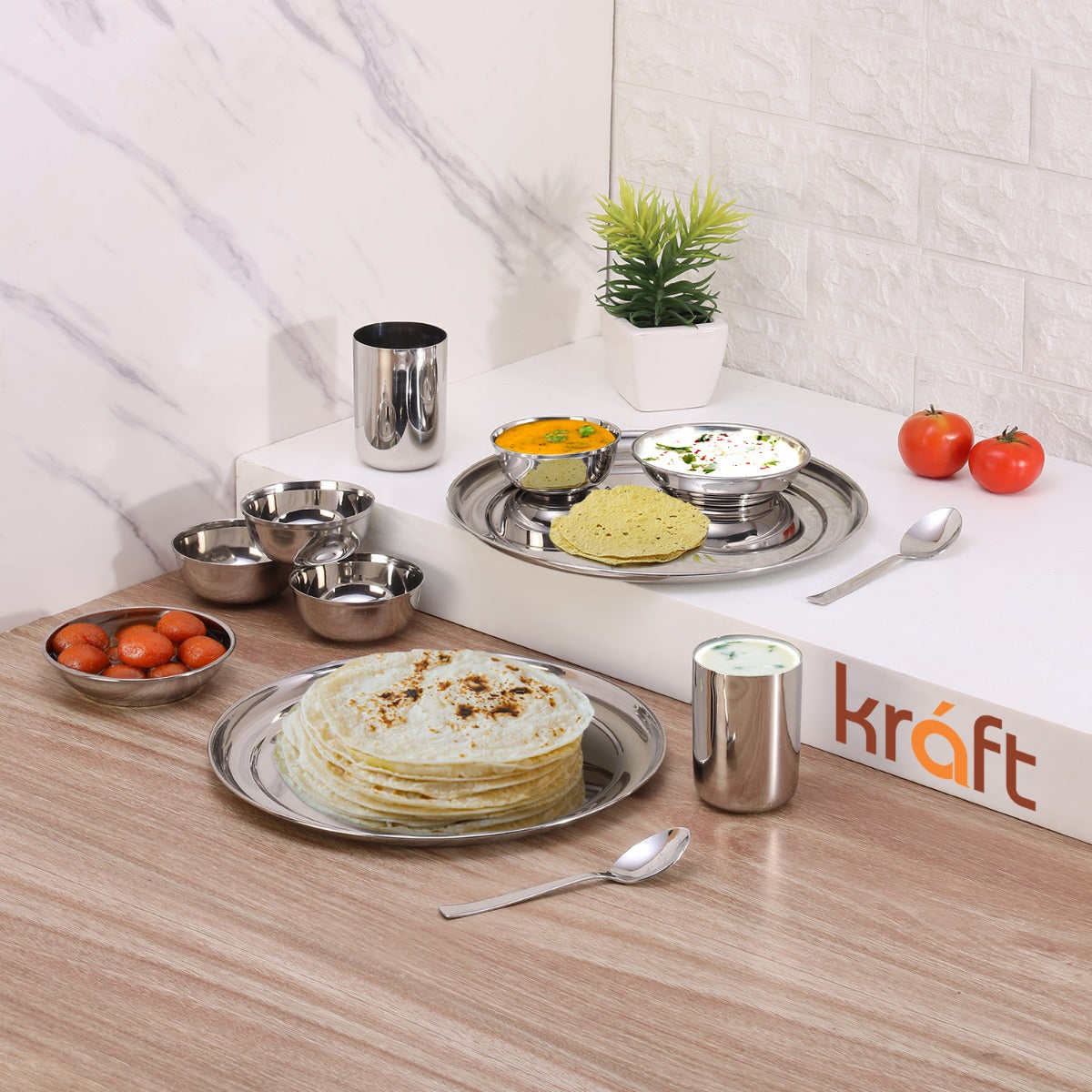 Stainless steel Princess dinner set 12-piece with plates, bowls, tumbler, and spoons, featuring Indian meal setup with roti, curry, sweets, and buttermilk in a modern kitchen setting